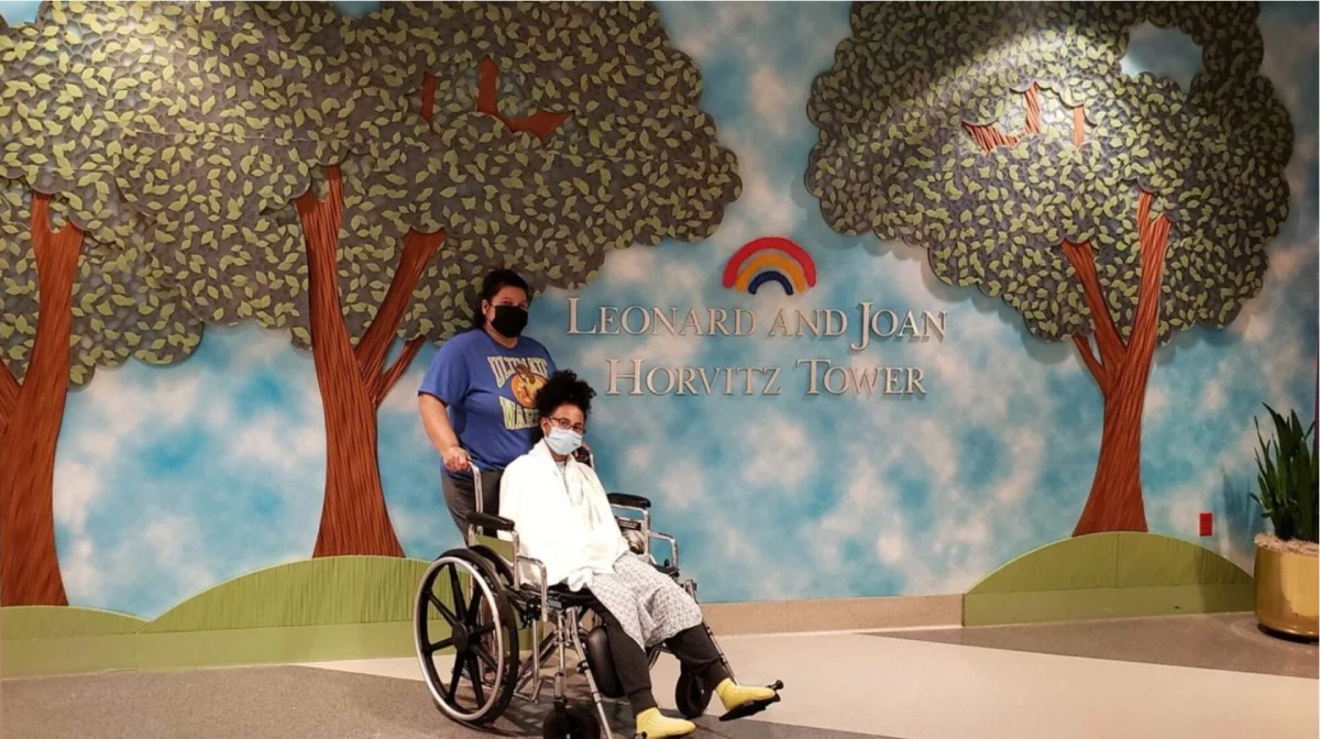 A person in a wheelchair and another standing beside them pose in front of a mural with trees and "Leonard and Joan Horvitz Tower" text. Both wear masks.