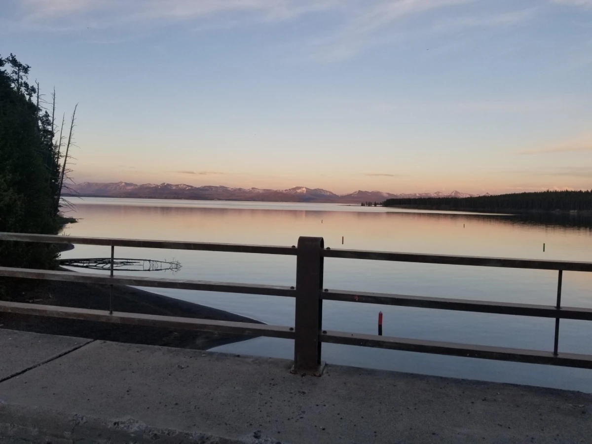 Calm lake at sunset with mountains in the distance, partially framed by a bridge railing. The sky is softly colored, creating a serene atmosphere.