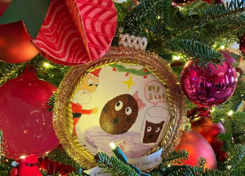 Close-up of a Christmas tree decoration featuring a child's drawing framed in gold. The colorful drawing depicts animated cookies and milk.