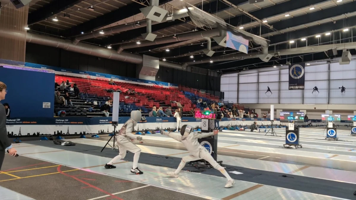 Two fencers in white suits and masks compete intensely on a brightly lit indoor strip. The arena, with red and blue seats, is partially filled with spectators, creating an energetic atmosphere.