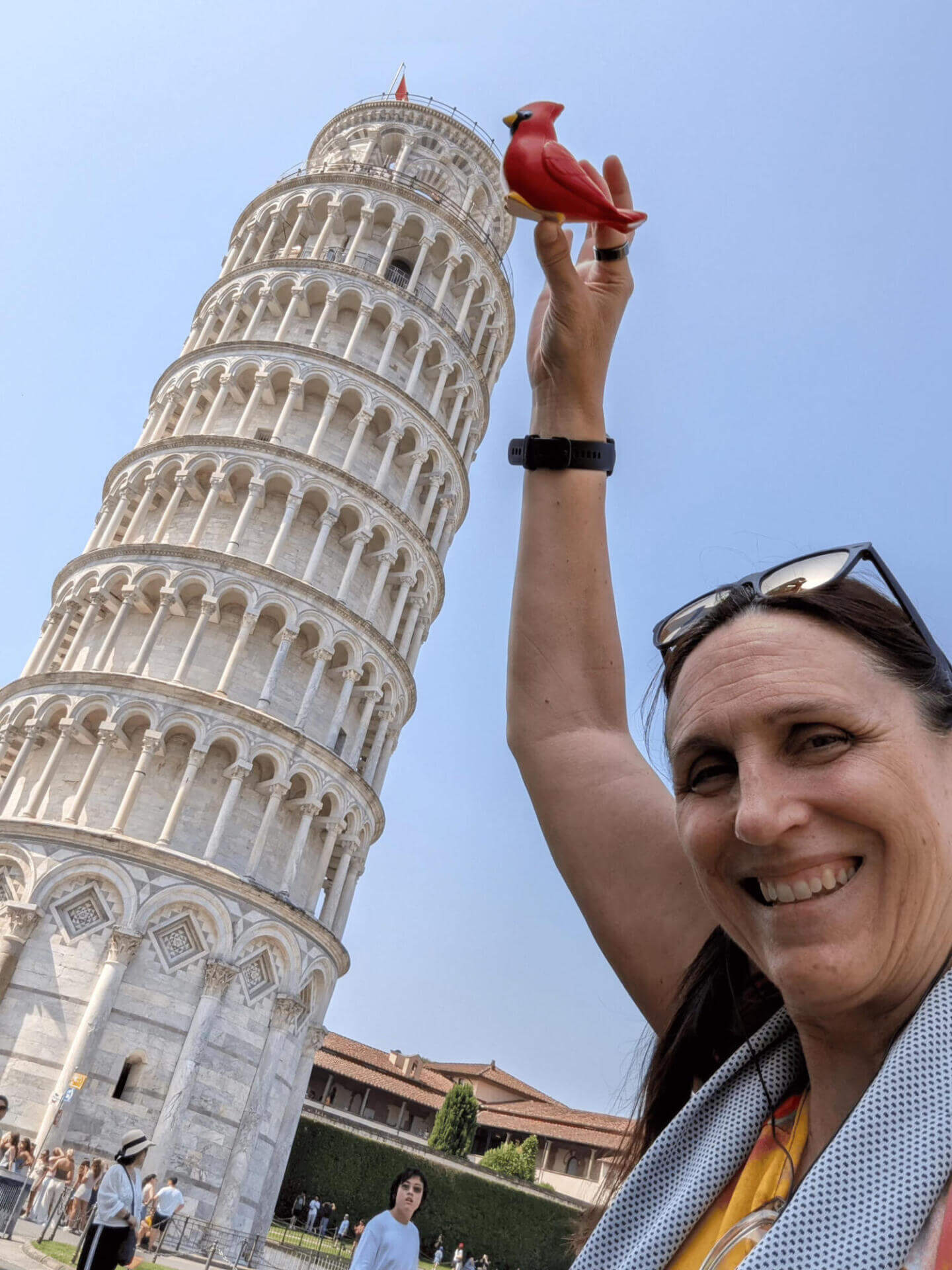 A woman smiling and holding up a small red cardinal figurine so it appears to rest on the Leaning Tower of Pisa in the background. The tower tilts noticeably against the clear blue sky, with other tourists visible at the base.