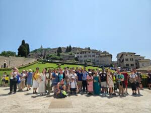 A large group of travelers posing together outdoors in front of a hillside town with stone buildings, green lawns, and tall cypress trees under a clear blue sky. The group stands closely together, smiling, with some people wearing hats, sunglasses, or backpacks.