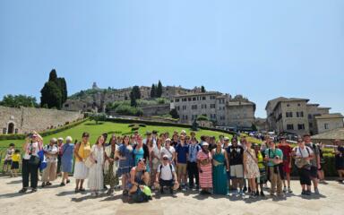 A large group of travelers posing together outdoors in front of a hillside town with stone buildings, green lawns, and tall cypress trees under a clear blue sky. The group stands closely together, smiling, with some people wearing hats, sunglasses, or backpacks.