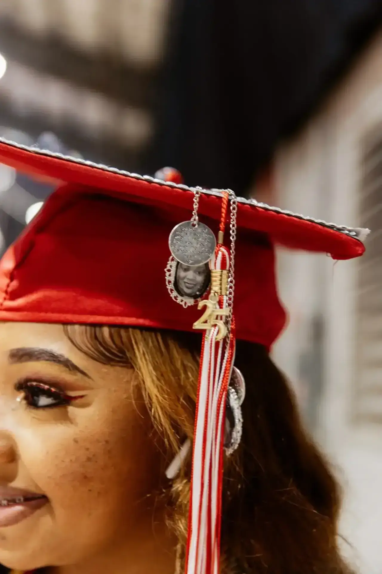 A graduate in a red cap with a "23" tassel smiles softly. The cap has a pendant featuring a person's photo, creating a celebratory and sentimental tone.