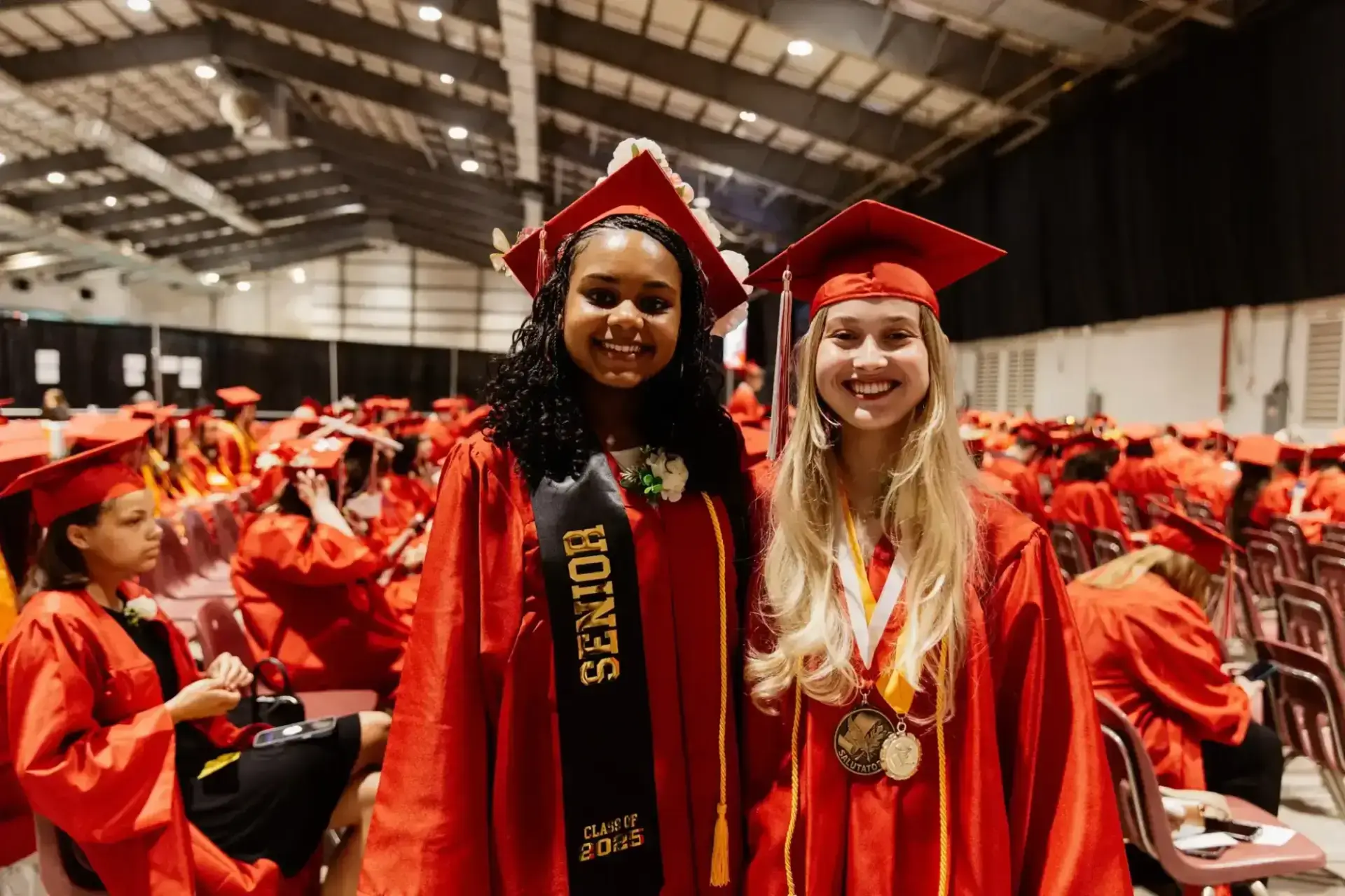 Two students in red caps and gowns smile at a graduation ceremony. One wears a "Senior" sash, while the other has medals. The background shows seated graduates.