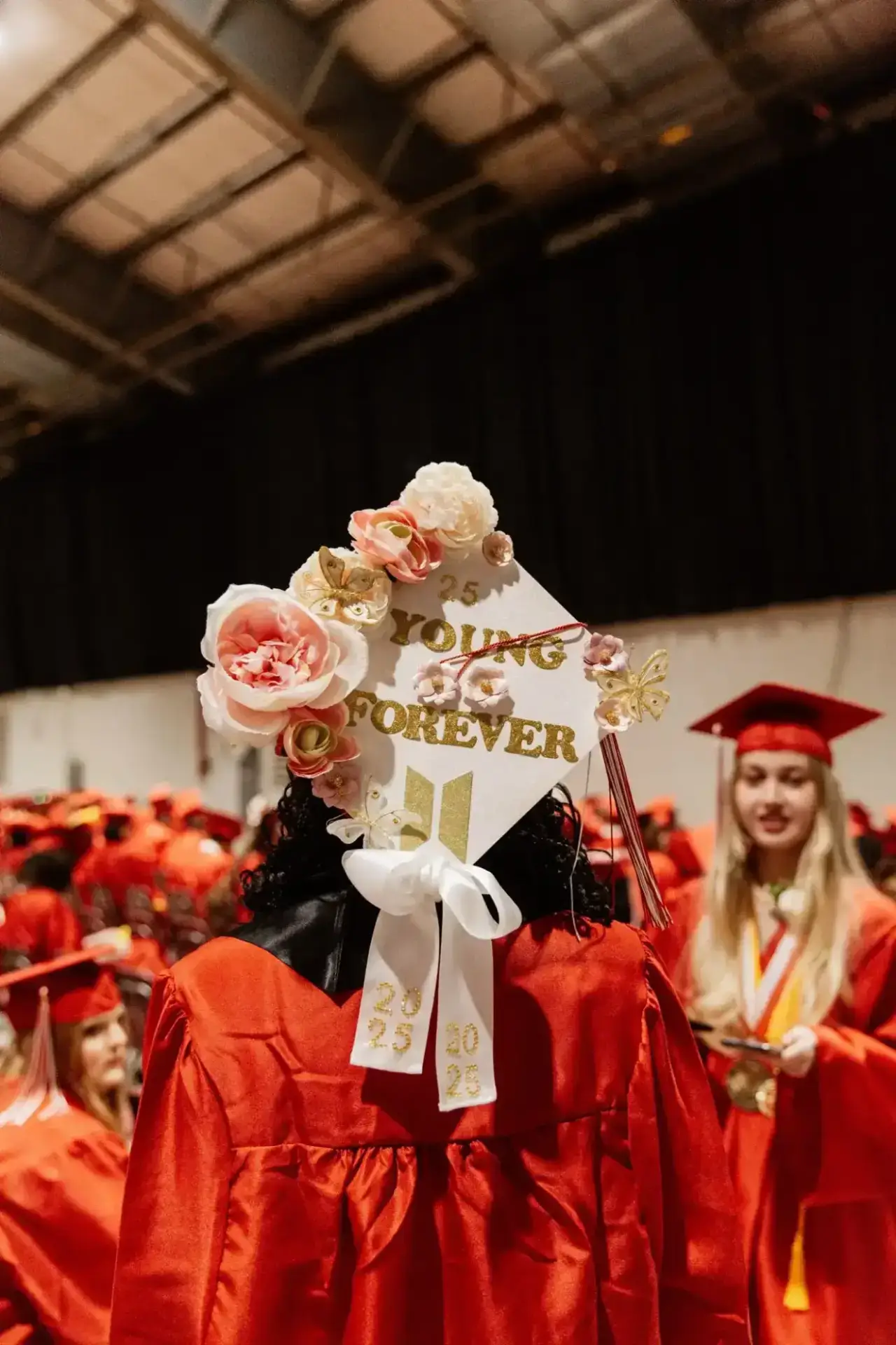 Graduate wearing a red gown and cap decorated with flowers and "Young Forever." Surrounded by other graduates, conveying celebration and achievement.