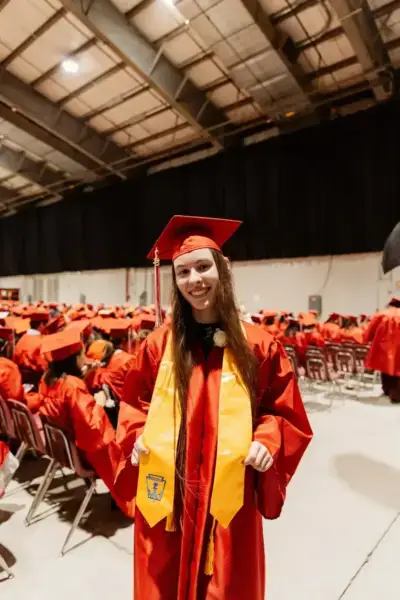 A graduate in red cap and gown stands smiling, wearing a yellow stole. She is surrounded by seated graduates in a large auditorium, conveying joy and achievement.