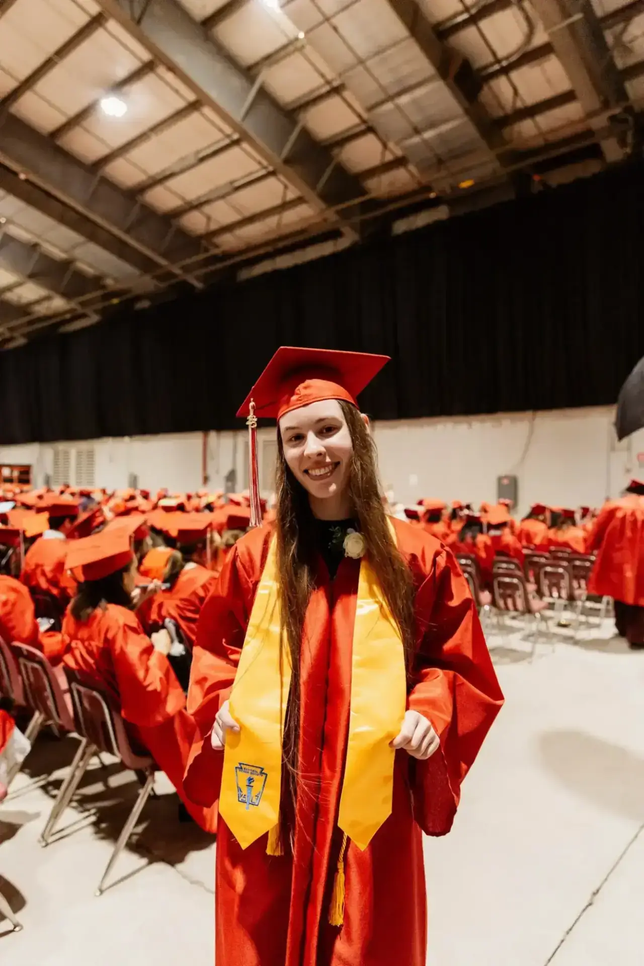 A graduate in red cap and gown stands smiling, wearing a yellow stole. She is surrounded by seated graduates in a large auditorium, conveying joy and achievement.