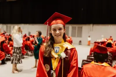 A young woman in a red graduation gown and cap stands smiling. She wears a yellow stole and medals. Behind her, graduates and guests mingle, creating a joyful atmosphere.