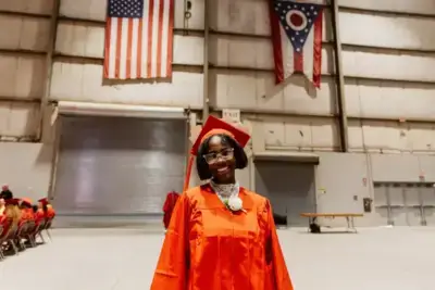 Smiling graduate in red cap and gown poses indoors, under US and Ohio flags, conveying pride and achievement. Rows of seated people in background.