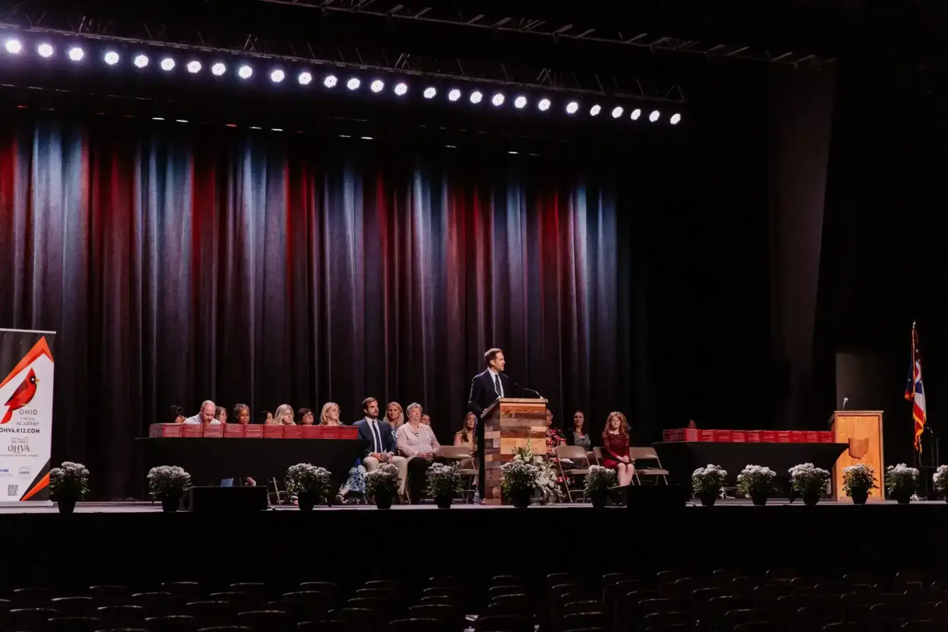 A man stands at a podium on a large stage with a curtain backdrop, addressing an audience. Seated people and potted plants line the stage. The atmosphere is formal.