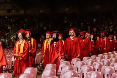 Graduates in red gowns and caps walk down an aisle between rows of empty seats during a ceremony.