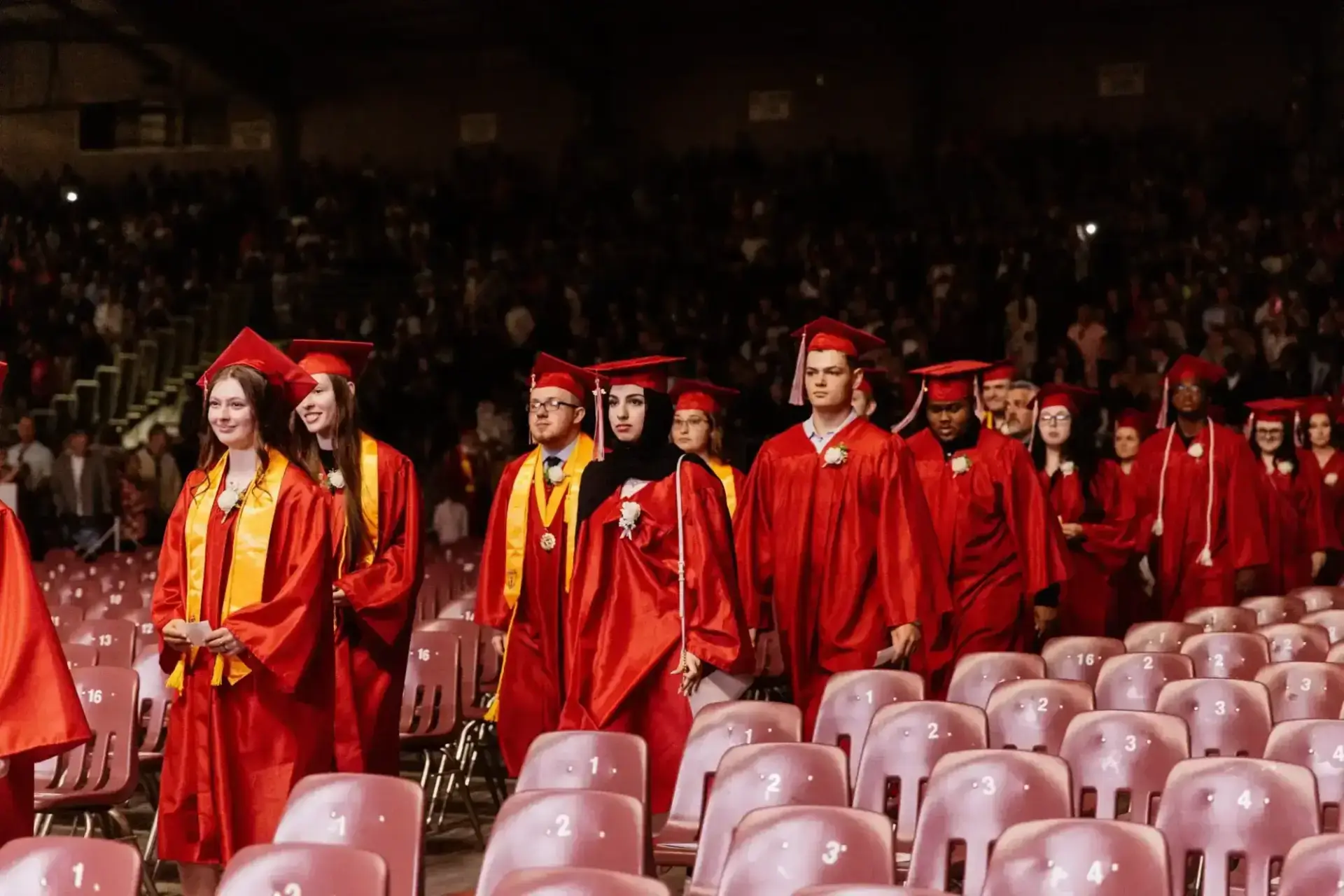 Graduates in red gowns and caps walk down an aisle between rows of empty seats during a ceremony.