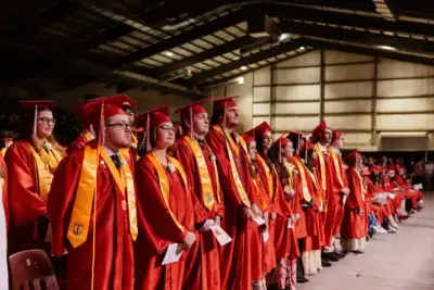 A group of graduates in red caps and gowns stand in a row indoors, looking forward with a sense of anticipation and pride.