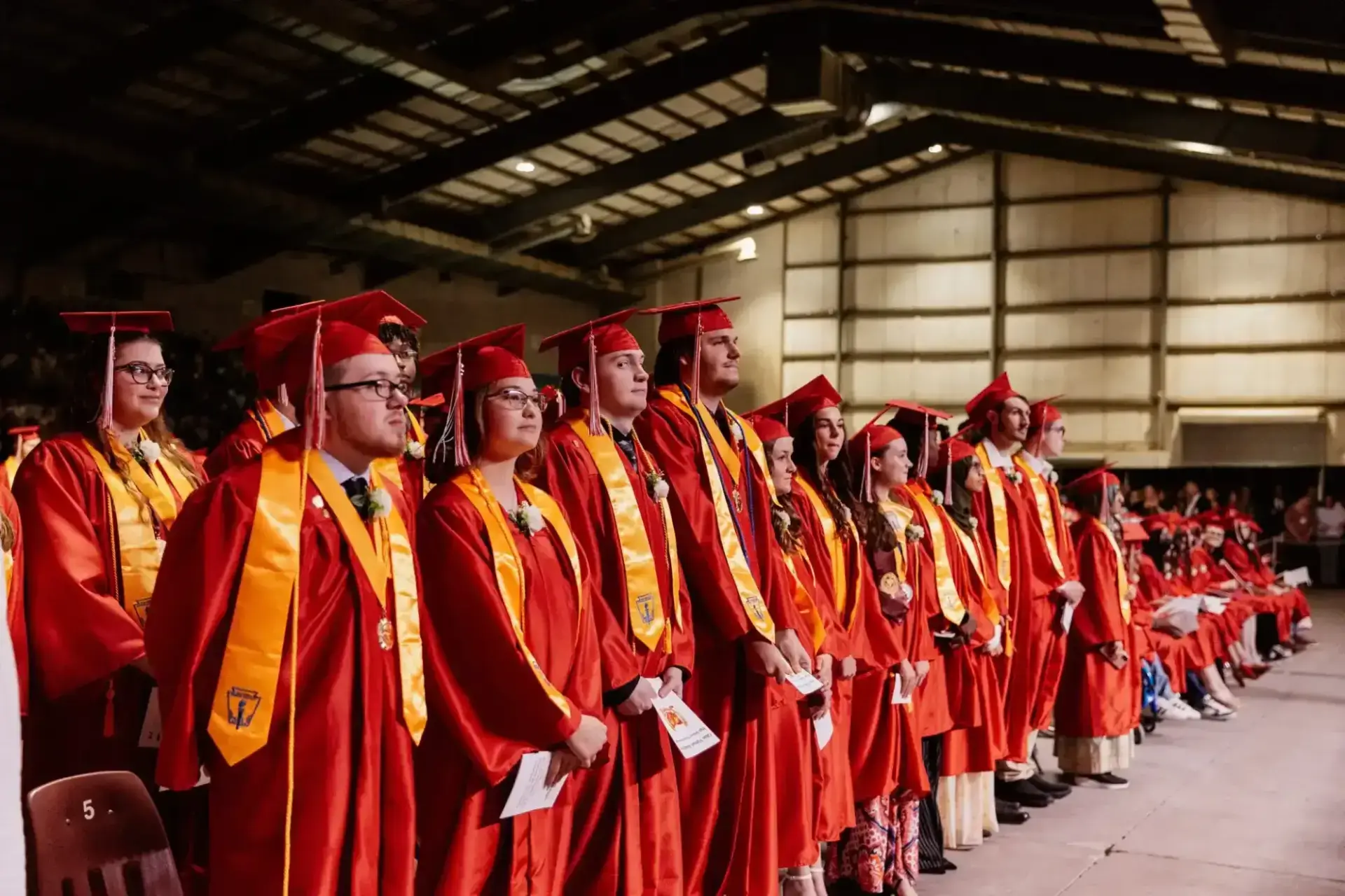 A group of graduates in red caps and gowns stand in a row indoors, looking forward with a sense of anticipation and pride.
