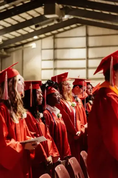 A row of graduates in red caps and gowns stands in a well-lit auditorium.