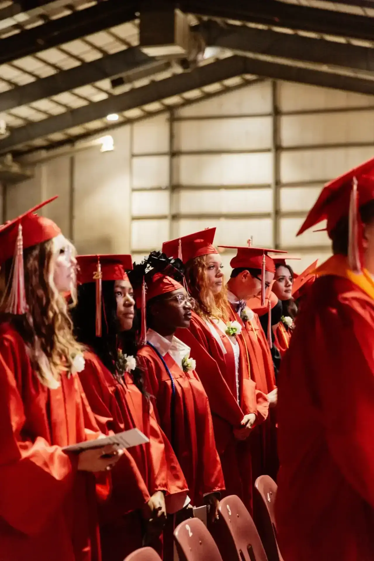 A row of graduates in red caps and gowns stands in a well-lit auditorium.
