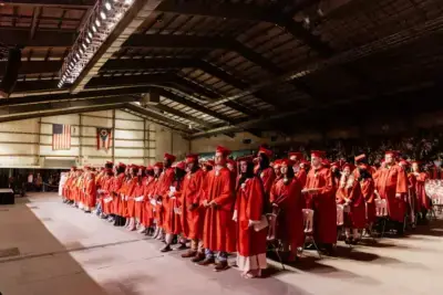 Graduates in red gowns and caps stand solemnly in rows during a ceremony in a large hall. U.S. and Ohio flags hang prominently in the background.