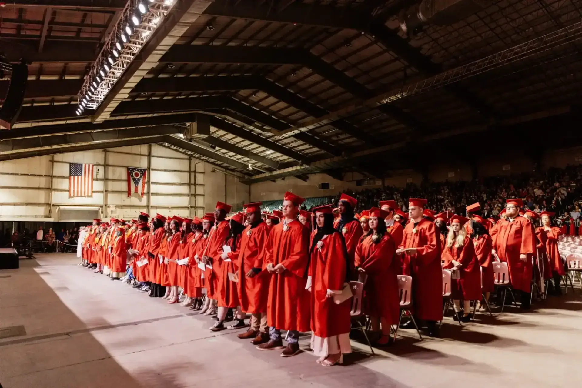 Graduates in red gowns and caps stand solemnly in rows during a ceremony in a large hall. U.S. and Ohio flags hang prominently in the background.