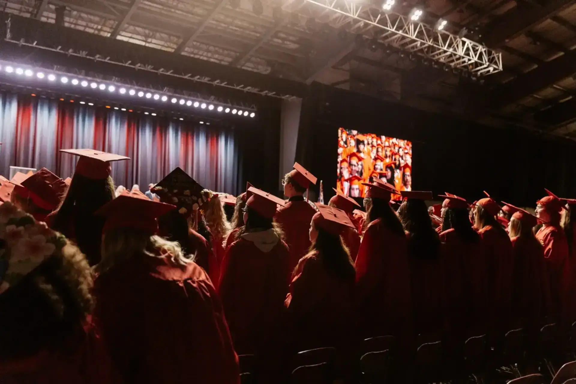 Graduates in red caps and gowns face a lit stage, with excitement and anticipation.