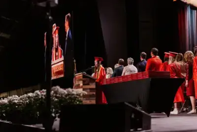 Graduation ceremony with a student in a red cap and gown speaking at a podium.