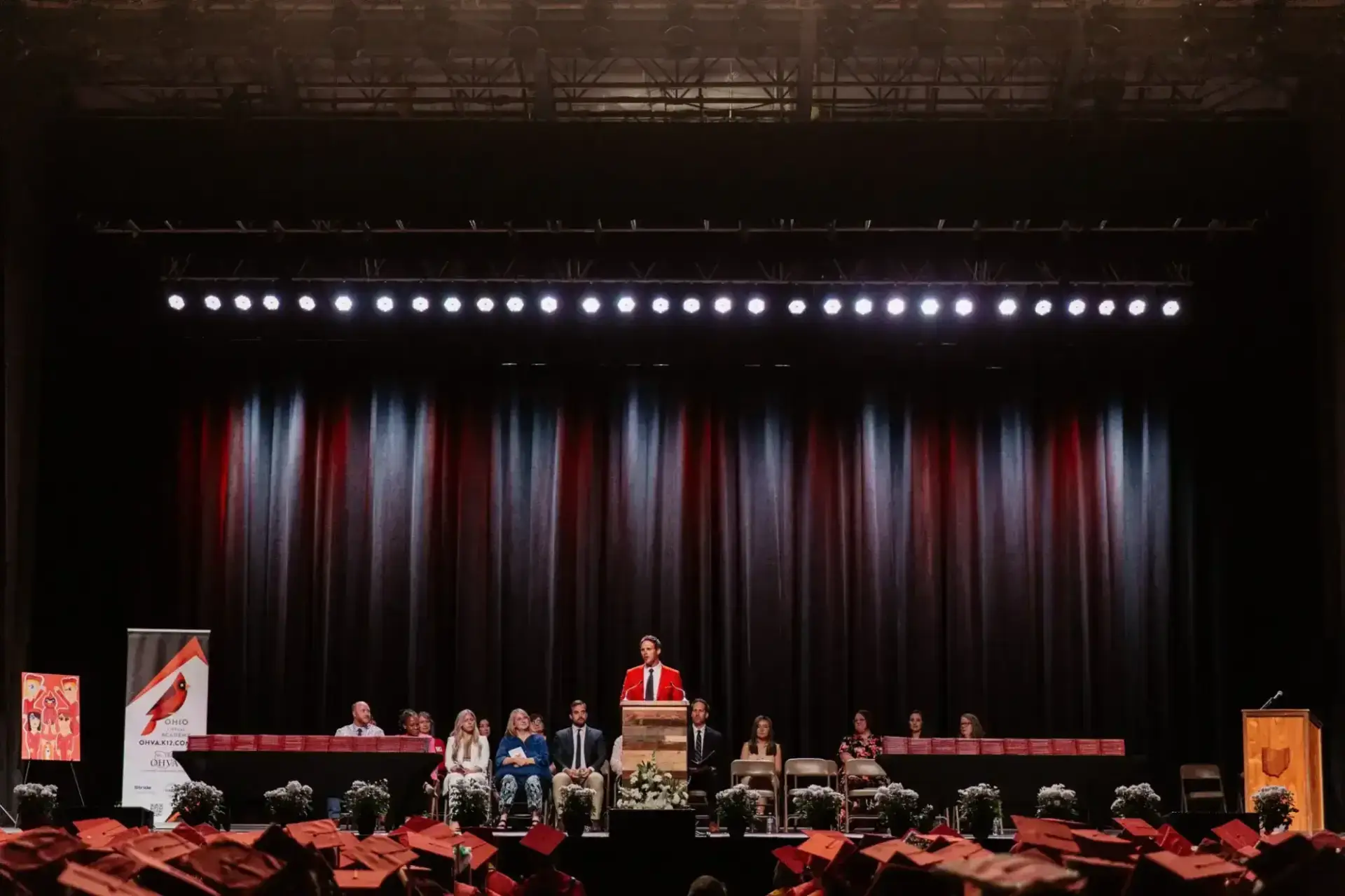A speaker in a red robe stands at a podium on a stage, with an audience of graduates in red caps watching.