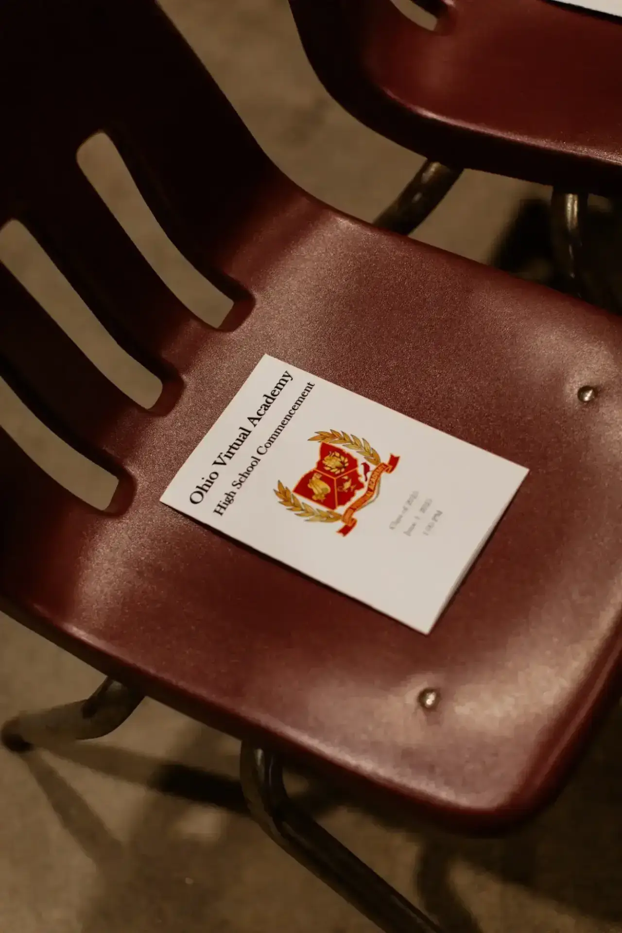 A maroon plastic chair with a high school commencement program on the seat. The cover features a crest, giving a formal and celebratory tone.