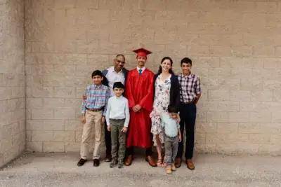 A joyful family of seven poses against a brick wall. A graduate in a red cap and gown stands in the center, surrounded by smiling relatives.