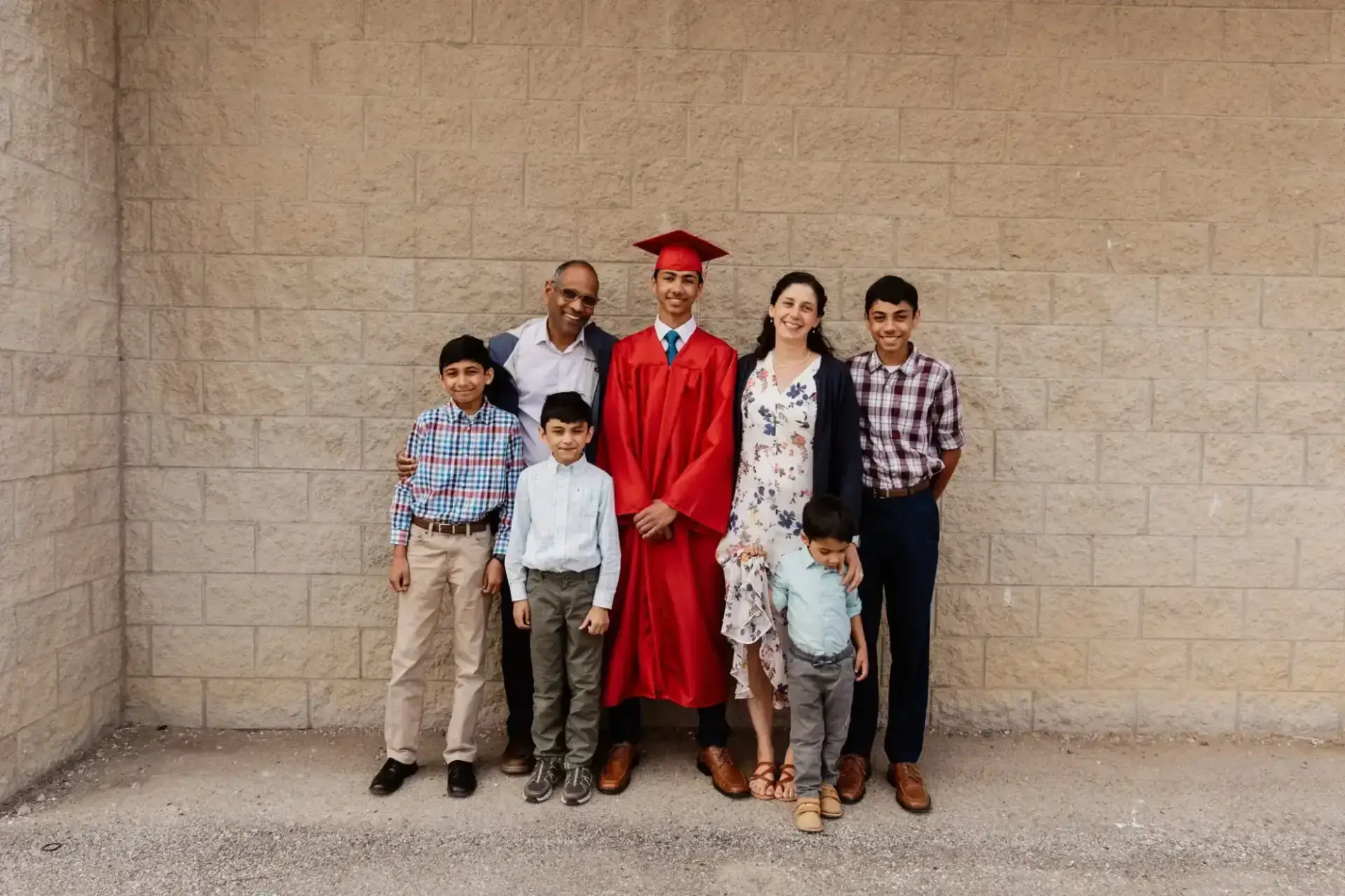 A joyful family of seven poses against a brick wall. A graduate in a red cap and gown stands in the center, surrounded by smiling relatives.