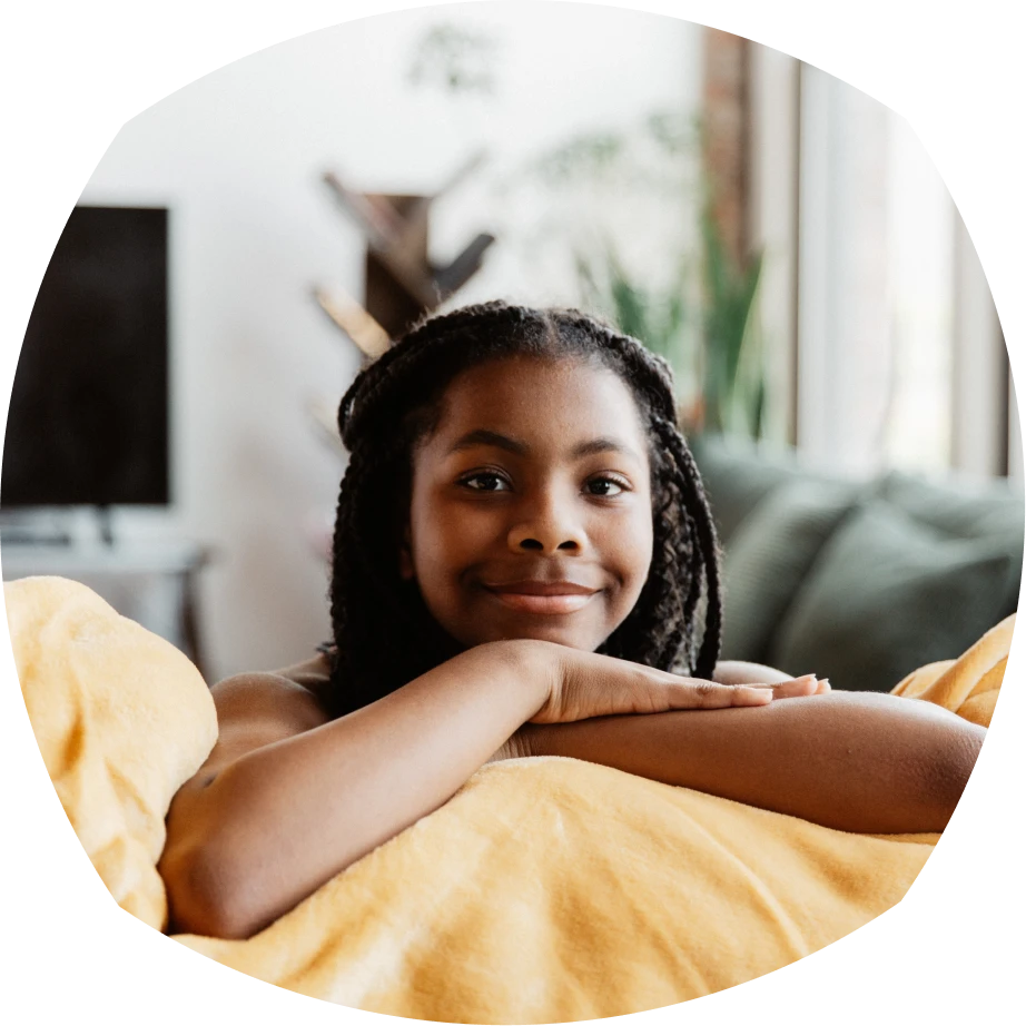 A young girl with braided hair leans on a couch, smiling warmly. She is wrapped in a yellow blanket, with a cozy living room in the background.