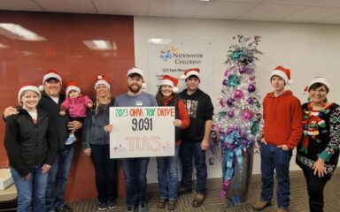 A group of smiling people wearing Santa hats stand in a brightly lit room holding a sign reading "2023 OHVA Toy Drive 9,091 TOYS" beside a festive Christmas tree adorned with pink and blue decorations.
