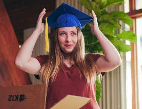 A woman with long hair smiles while adjusting a blue graduation cap with a yellow tassel. She's indoors, near a large plant, creating a celebratory mood.