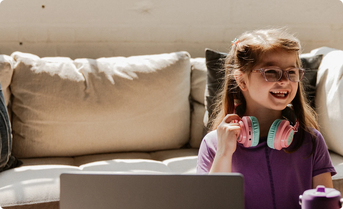Smiling young girl with glasses and colorful headphones sits in front of a laptop in a sunlit room, exuding happiness and a sense of comfort.