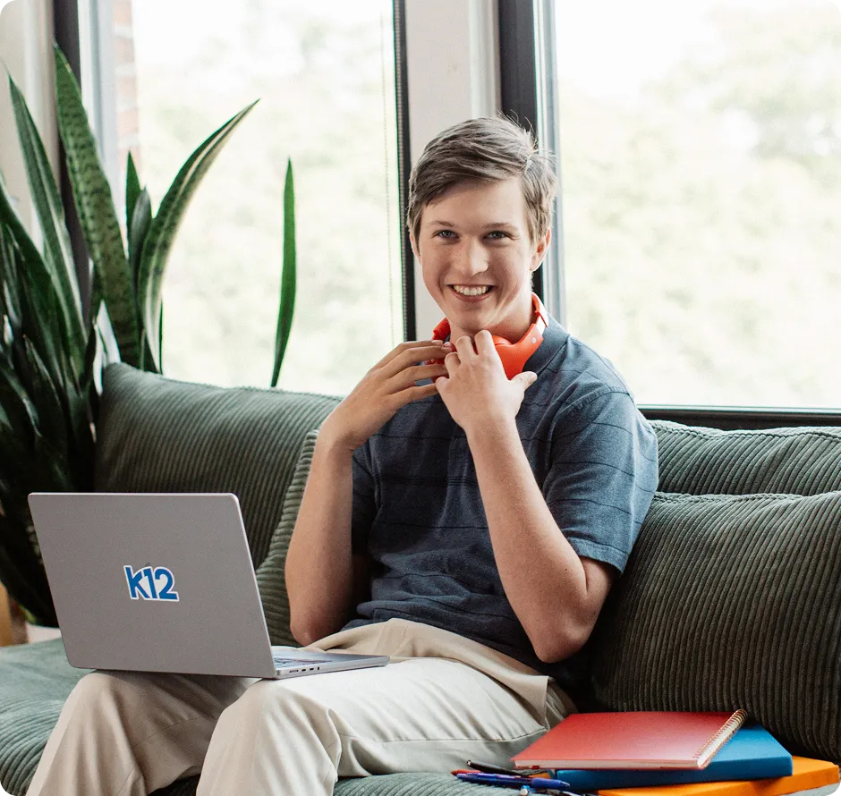 Smiling person sitting on a couch with a laptop marked "k12." They wear headphones and have notebooks nearby, conveying a studious, relaxed mood.