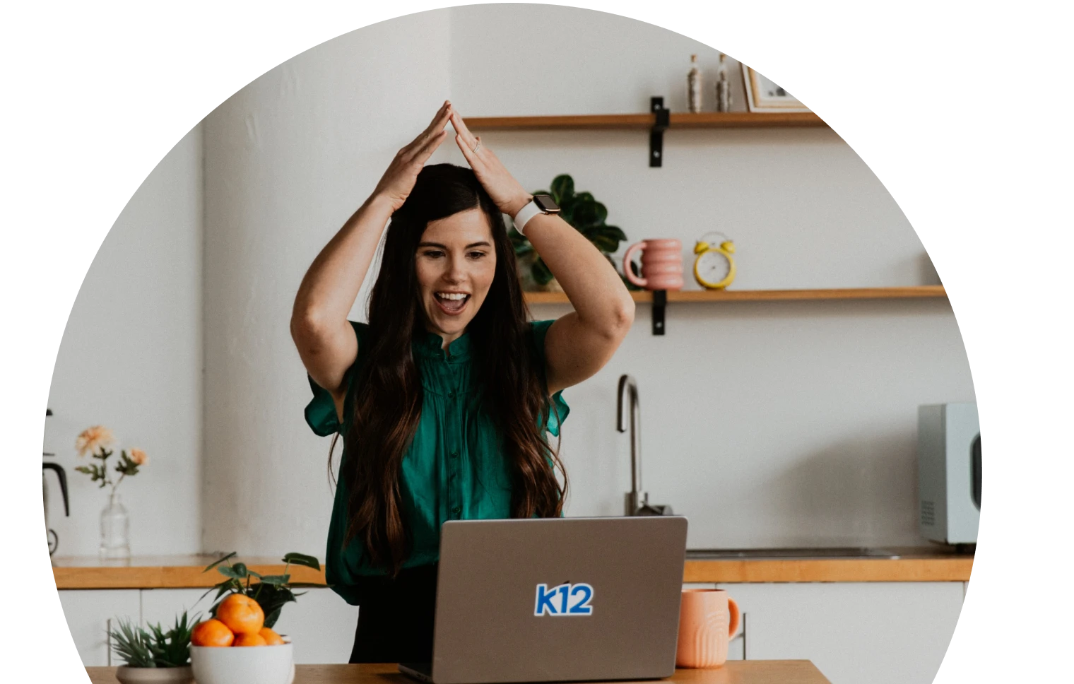 A woman in a teal blouse happily gestures with excitement in front of a laptop. The background is a cozy kitchen with shelves, plants, and vibrant decor.